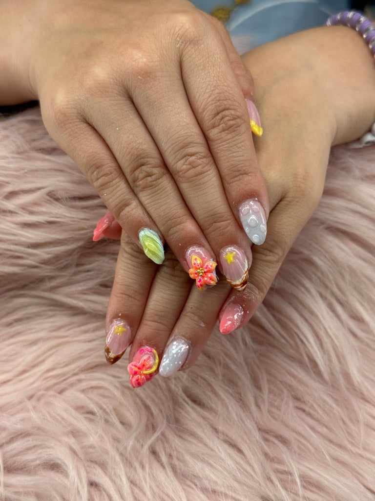 Hands with colorful 3D nail art featuring flowers, glitter, and pastel designs on a fluffy surface